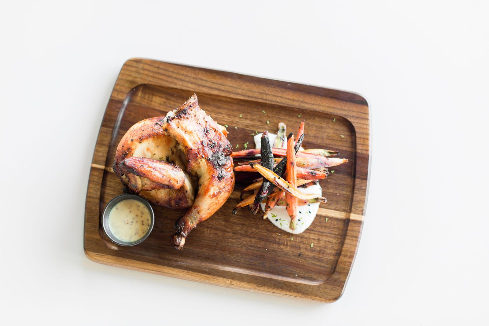 Half a rotisserie chicken with roasted rainbow carrots and a ramekin of dressing on a walnut board