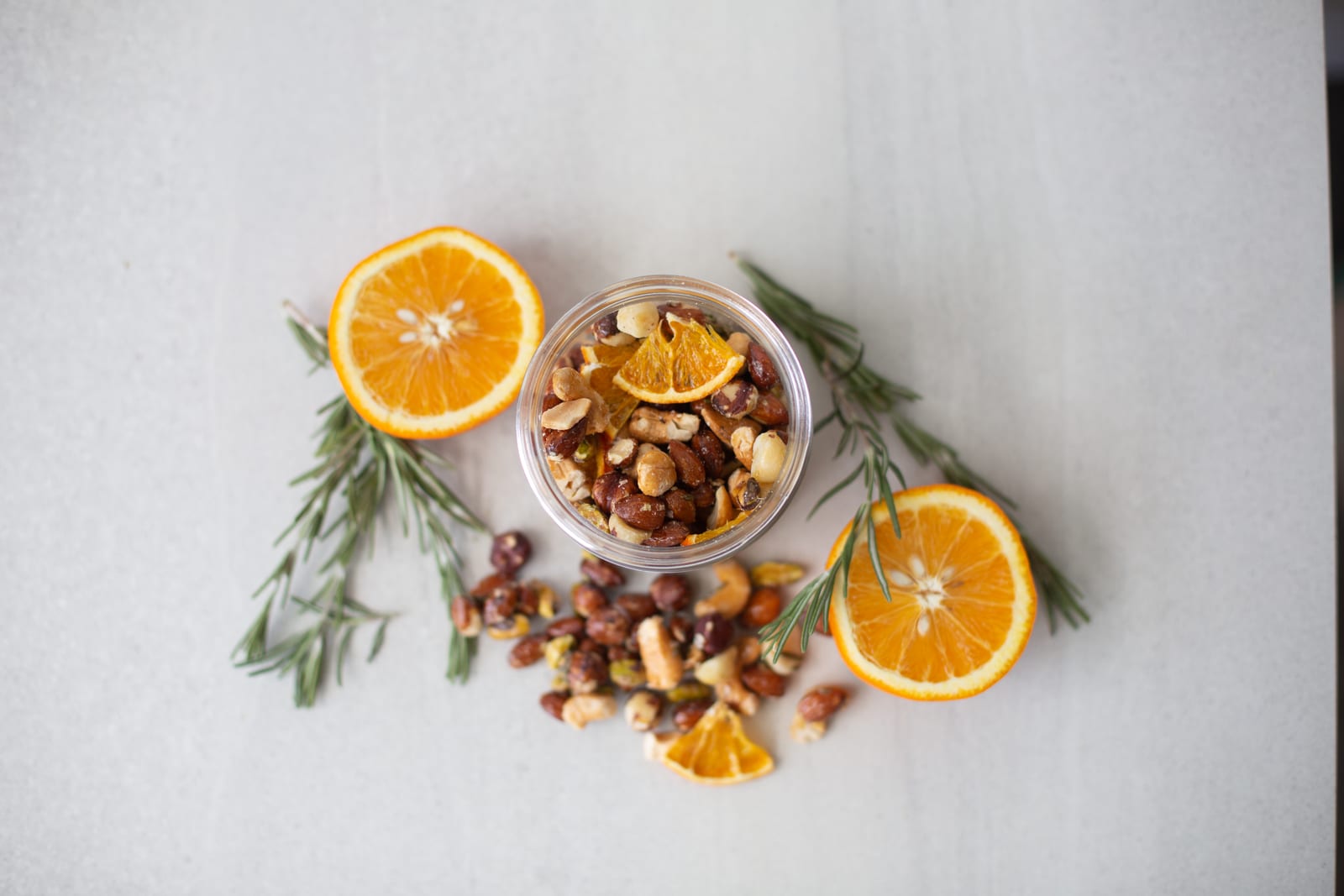 Spiced orange nuts in a jar, with the mix spilled beside it among fresh orange slices and rosemary