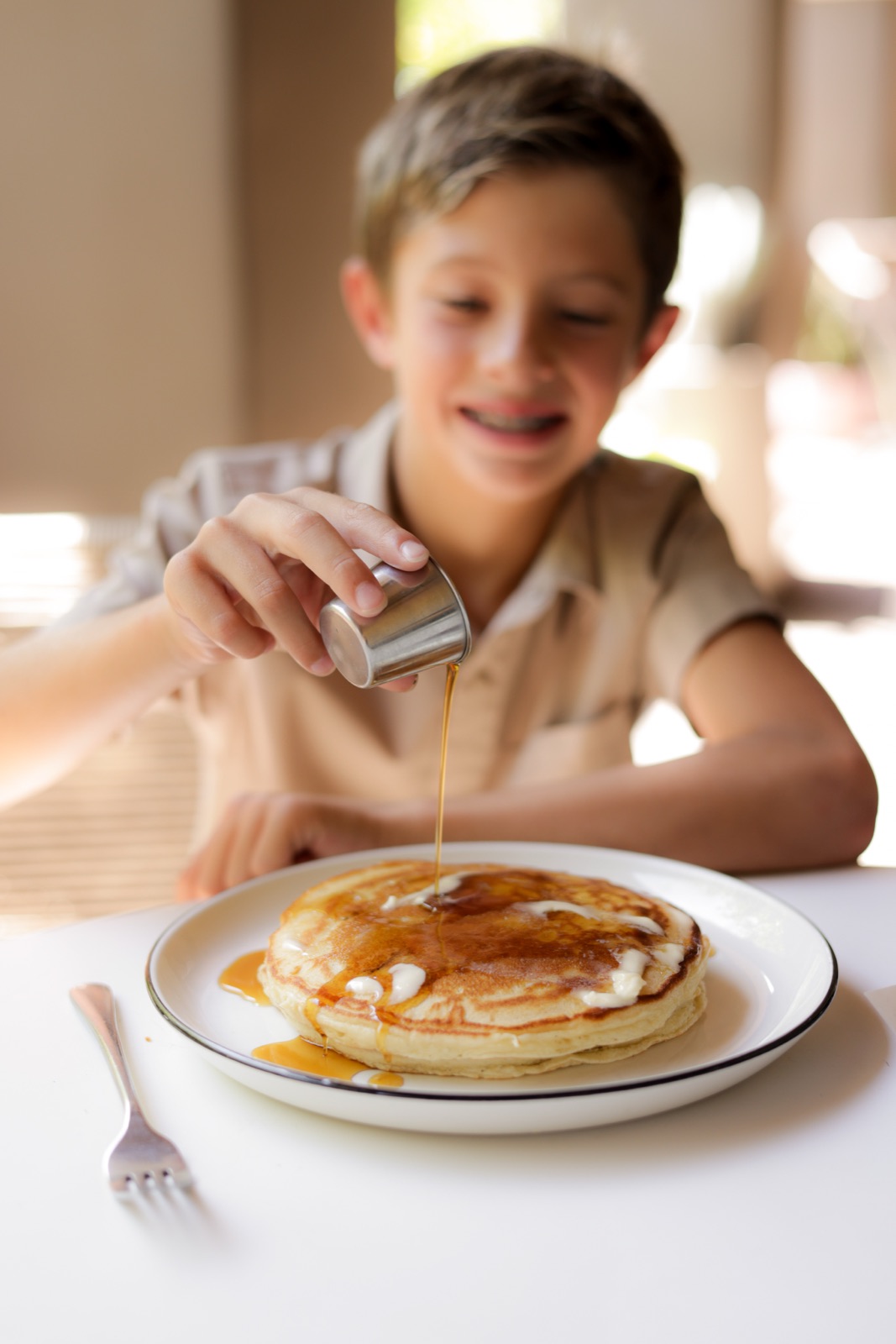 A boy pouring syrup over a stack of pancakes in the morning light