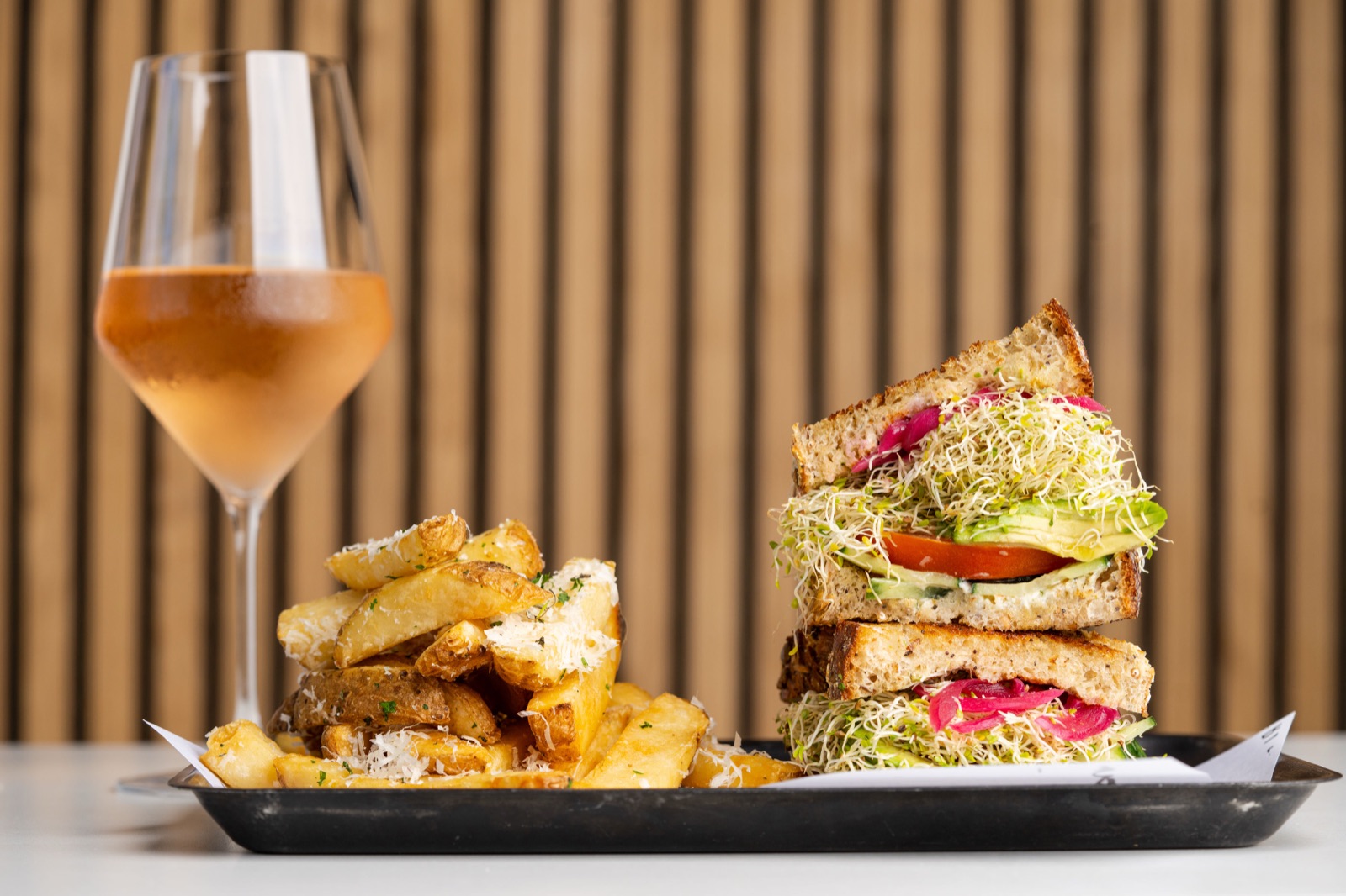 A veggie sandwich stacked with sprouts and avocado beside parmesan steak fries and a glass of rose