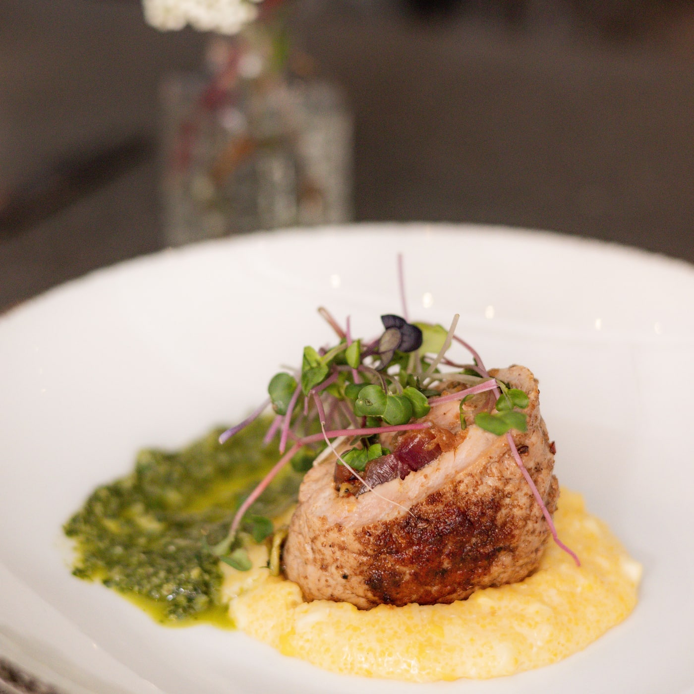 A plated course at the table: pork over soft polenta with salsa verde and micro greens, white flowers behind it