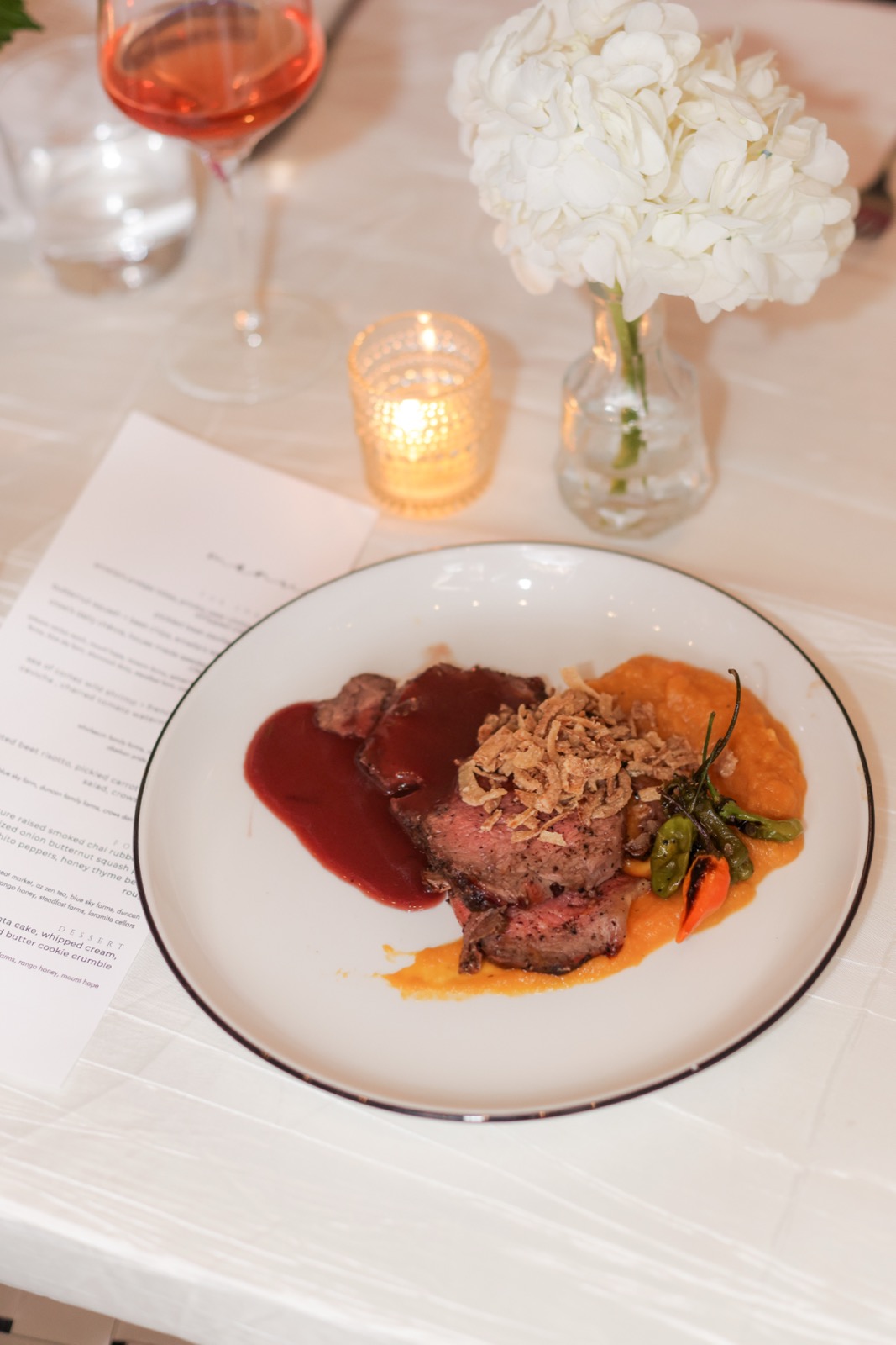 The printed menu for the evening beside a plated course, a candle and white flowers